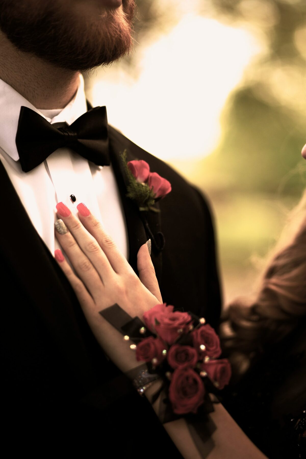 Close-up of a black tuxedo and bow tie with a rose boutonniere, a partner's hand and rose corsage resting on his chest