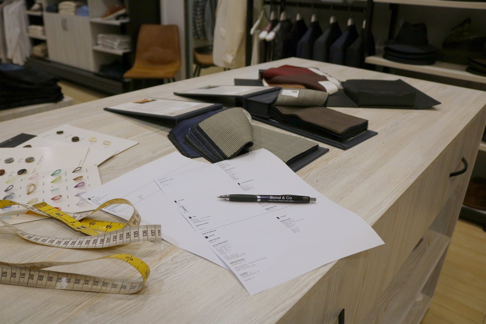 Fabric swatch books, an order sheet, a Bond & Co pen and a tape measure laid out on the tailoring counter in store