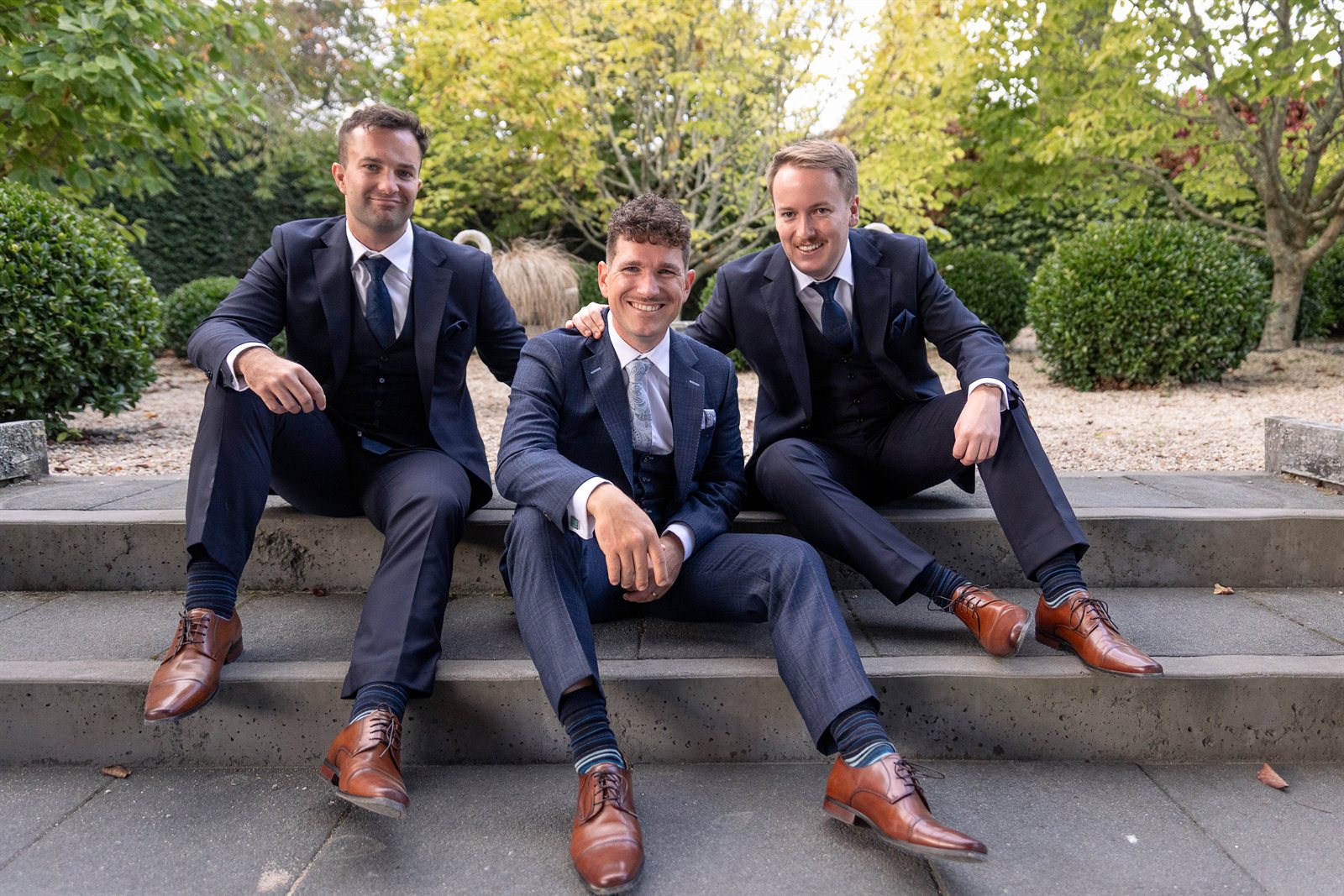 Three groomsmen in navy three-piece suits and brown brogues sitting together on stone garden steps
